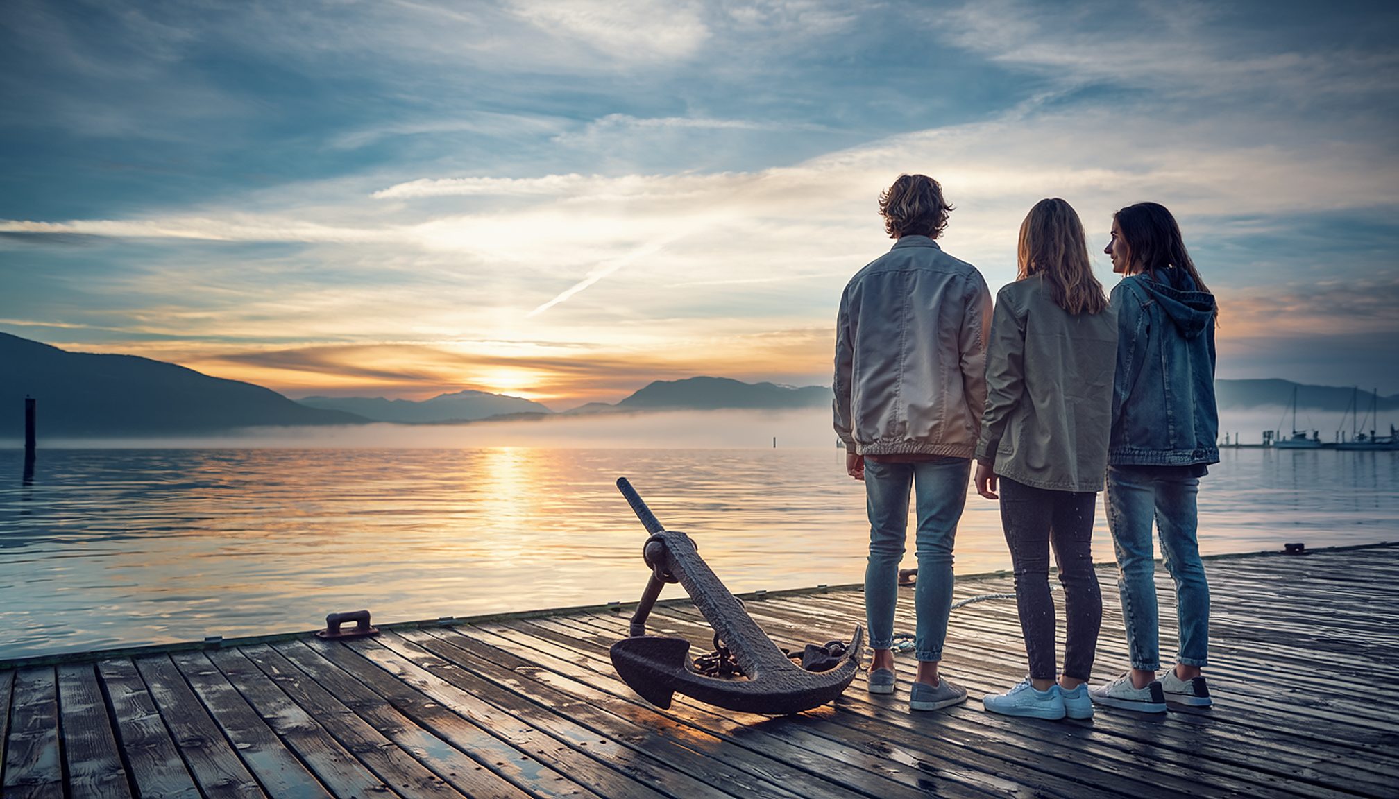 Three young adults standing together on a pier at sunrise beside an old iron anchor, looking out over a calm harbor.